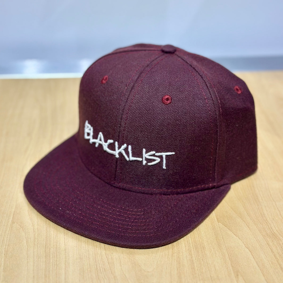 Maroon snapback cap with white 'BLACKL1ST' logo on front; flat brim.